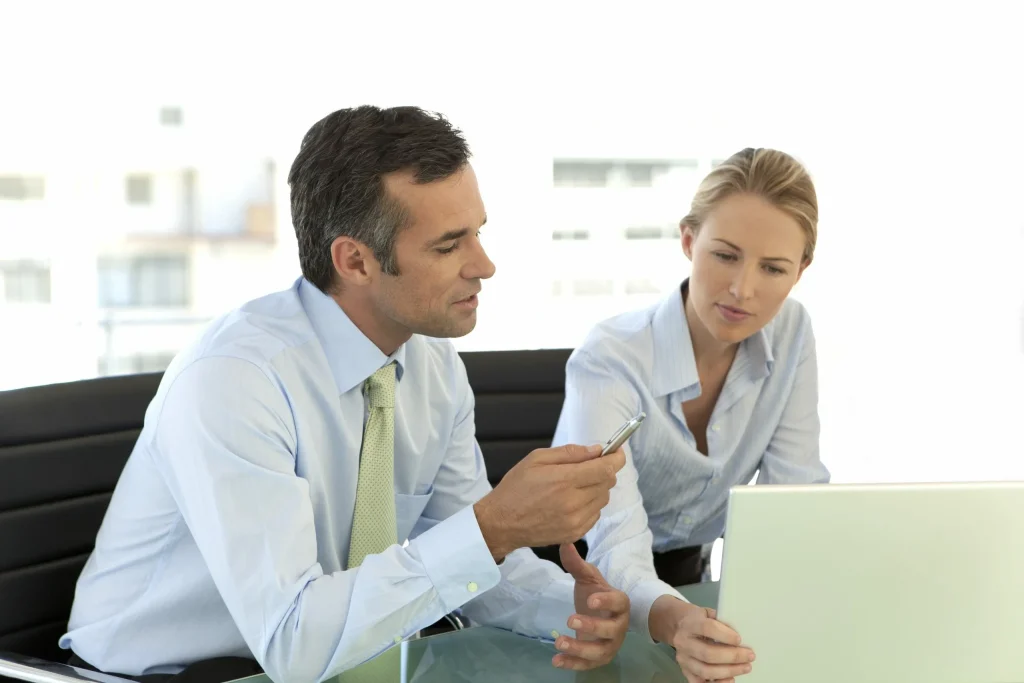 A man and woman in business attire discuss over a laptop in a bright office.