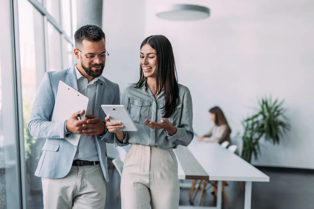 A man and woman in business attire laugh while looking at a tablet in a bright office.