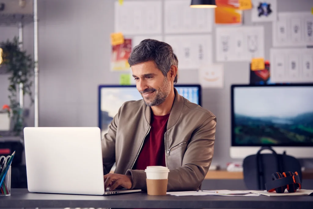 Man with graying hair in a casual jacket works on a laptop at a cluttered desk, surrounded by papers, a coffee cup, and a double-screen setup.