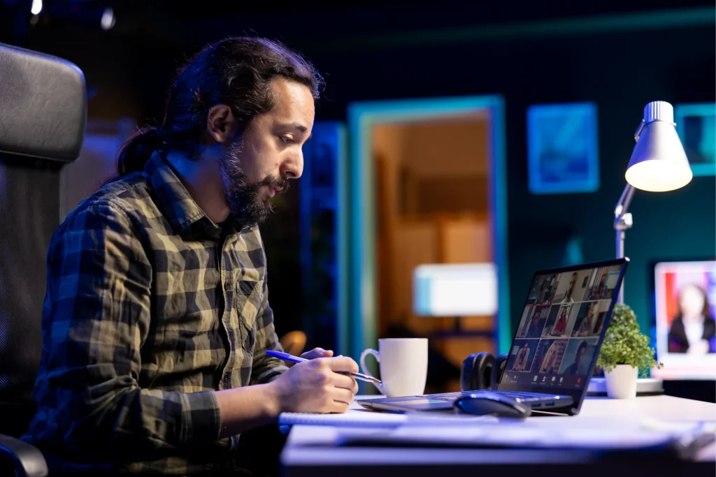 Man with long hair and a checkered shirt sits at a desk, focused on a laptop screen showing a video call. The room is dimly lit with a modern lamp.