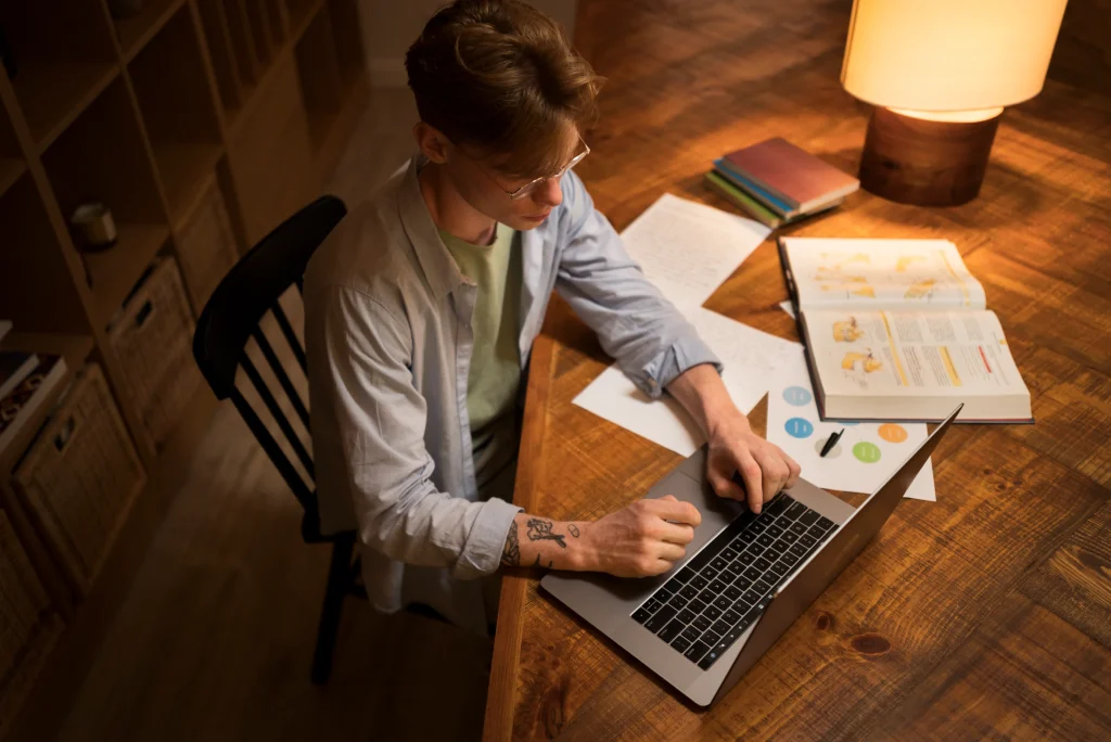 A person sits at a wooden desk using a laptop, surrounded by open books, notes, and a glowing lamp. The setting is cozy and focused.