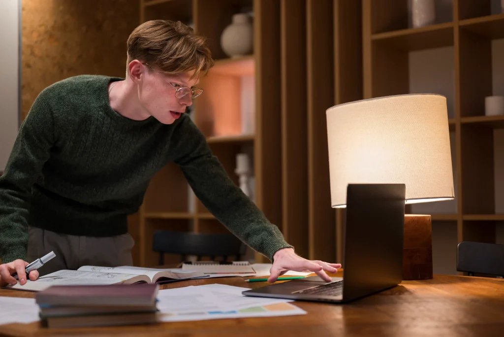 A young person in glasses and a sweater leans over a wooden table, intensely focusing on a laptop. Papers are scattered around, illuminated by a table lamp.