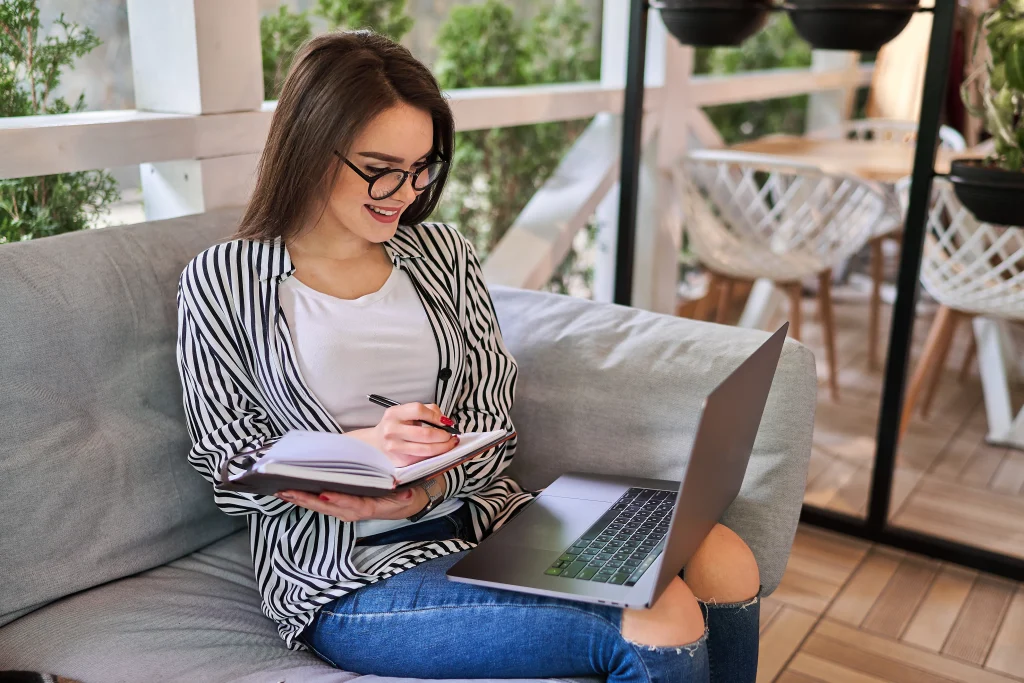 Woman with glasses writing in a notebook while smiling at a laptop. She sits on a cozy sofa near large windows, conveying a relaxed work vibe.