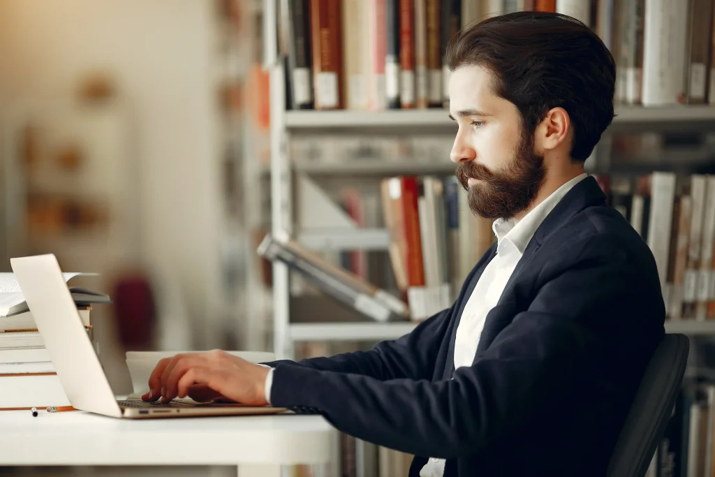 A man in a navy blazer concentrates on his laptop in a library. Bookshelves filled with books are in the background, creating a studious atmosphere.