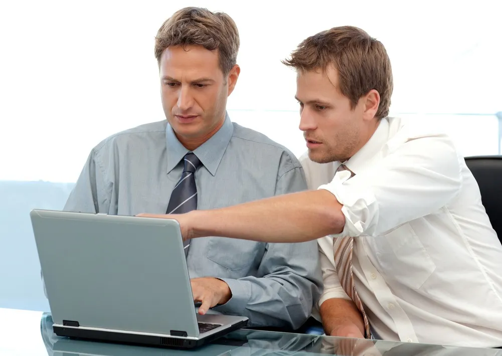 Two men in business attire work together at a laptop, one pointing at the screen, conveying collaboration and focus in a modern office setting.