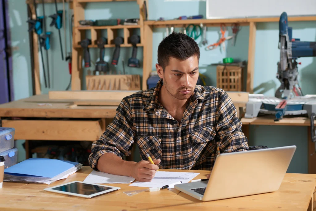 waist-up-shot-handsome-carpenter-planning-work-project-looking-laptop-with-pencil-hand